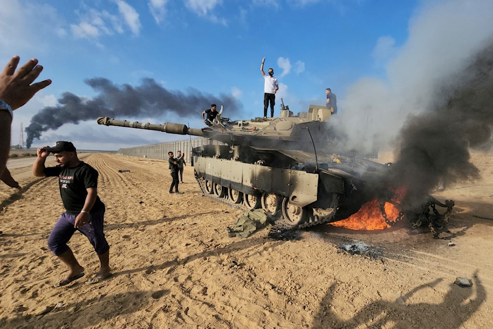 Palestinians celebrate by a destroyed Israeli tank at the Gaza Strip separation wall east of Khan Yunis, occupied Palestine, October 7, 2023 (AP)
