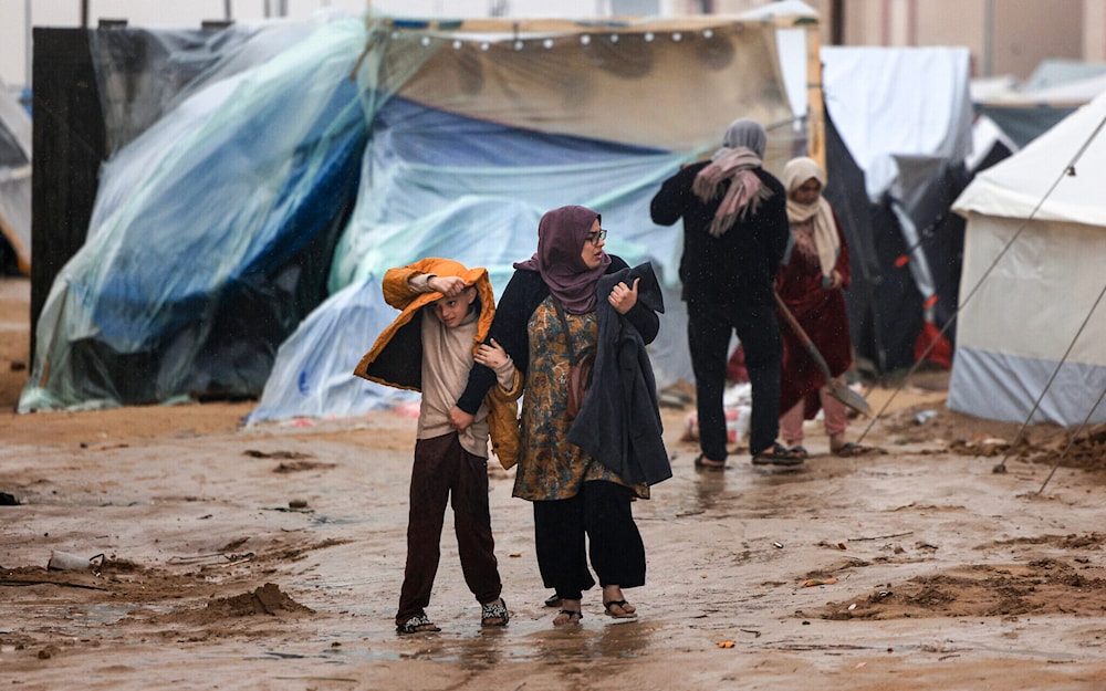 Palestinians walk under the rain at a camp for displaced people in Rafah, in the southern Gaza Strip where most civilians have taken refuge on December 13, 2023 (AP)