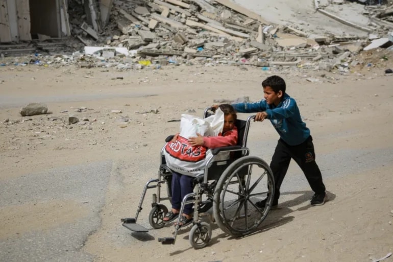A Palestinian child pushes his little sister in a wheelchair past a destroyed building in Gaza City amid the ongoing Israeli genocide on Gaza, March 28, 2024. (AFP)