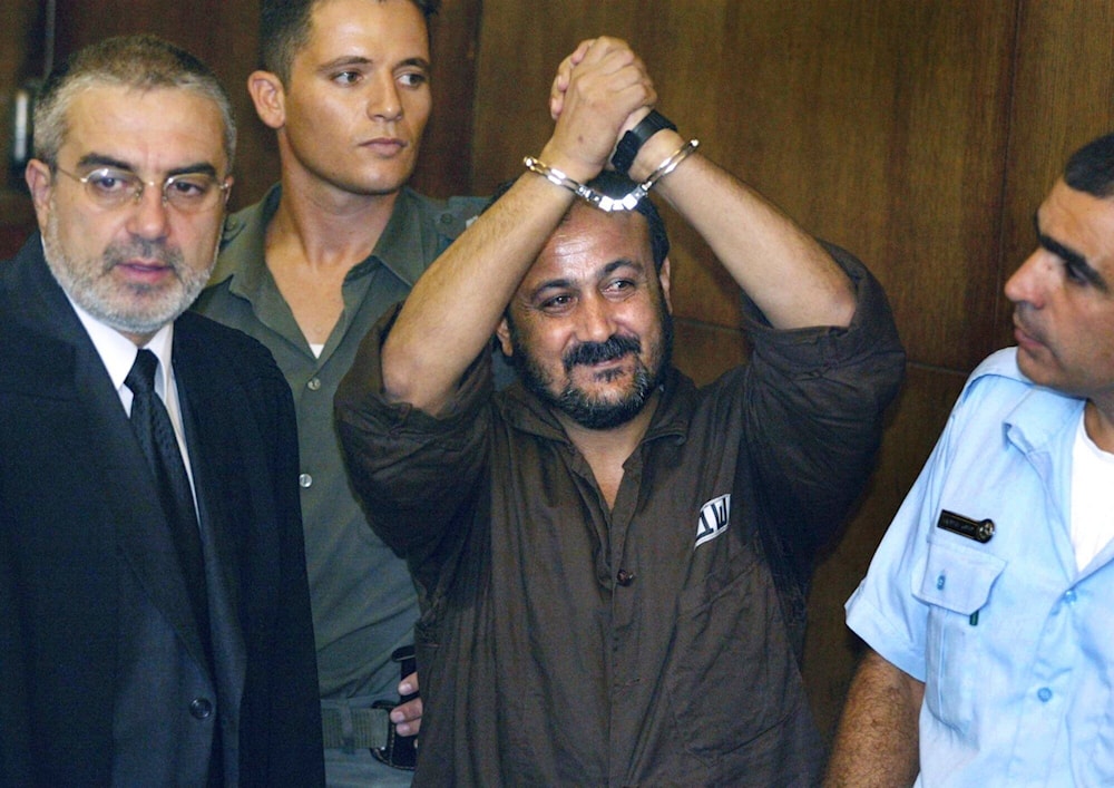 Marwan Barghouti, center, raises his handcuffed hands in the air on the opening day of his trial in 'Tel Aviv', occupied Palestine on Aug. 14, 2002.(AP)