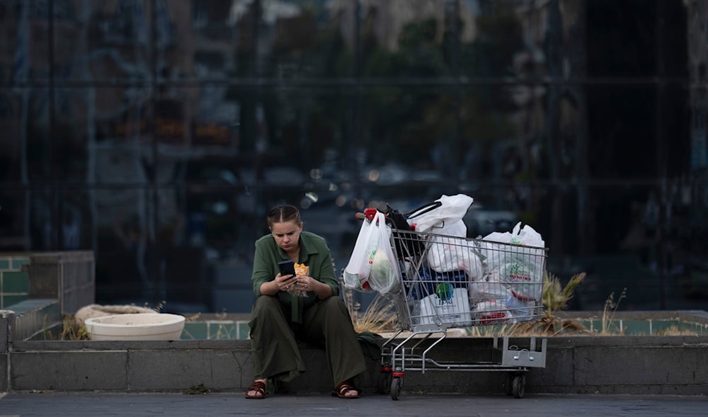 A woman looks at her phone while eating a sandwich next to a shopping cart in occupied Haifa, 'Israel', Thursday, Aug. 15, 2024. (AP)