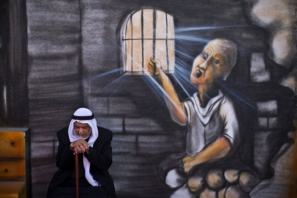 Palestinian refugee Shaher Alkhateeb, 73, who witnessed the Nakba, poses for a picture in front of a wall painted with a mural depicting a prisoner jailed in 