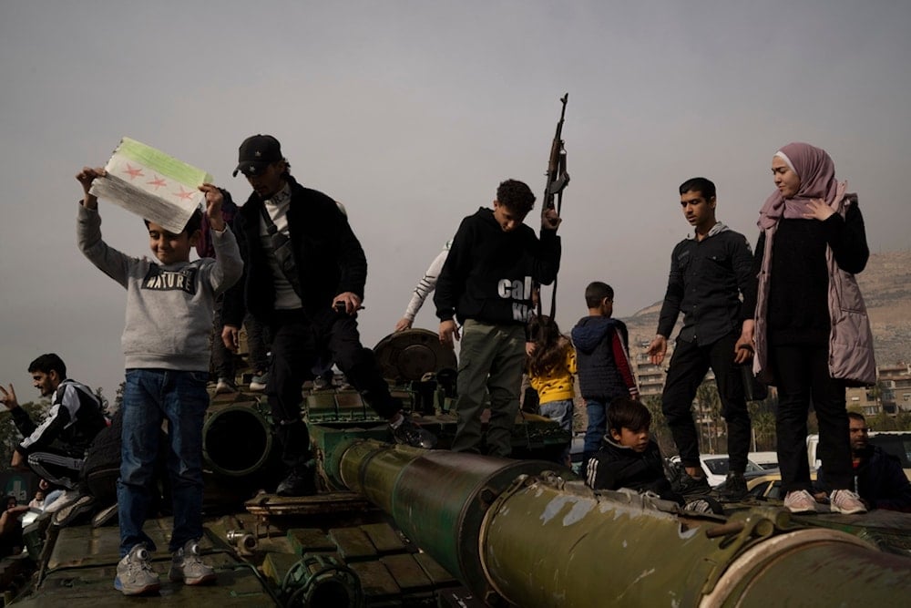 A youth holding a rifle borrowed from a Syrian opposition fighter poses for a picture on the top of a government forces tank that was left on a street, at the Umayyad Square in Damascus, Syria, Wednesday, December 11, 2024 (AP)