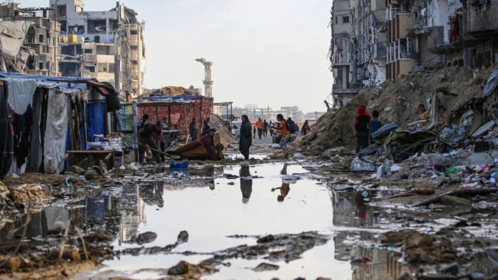 Forcibly displaced Palestinians walk around a puddle in front of destroyed buildings and tents in Khan Younis amid the Israeli genocide in Gaza. (AFP)