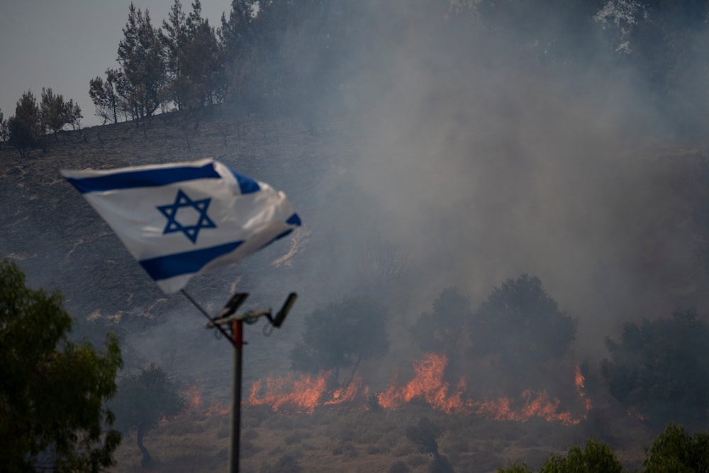 An Israeli flag flutters next to a fire burning in an area near the border with Lebanon, northern occupied Palestine in Safed, June 12, 2024 (AP)