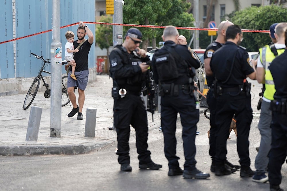 An Israeli settlers walks past Israeli occupation forces investigating the scene of a deadly explosion in Tel Aviv, occupied Palestine, July 19, 2024 (AP)