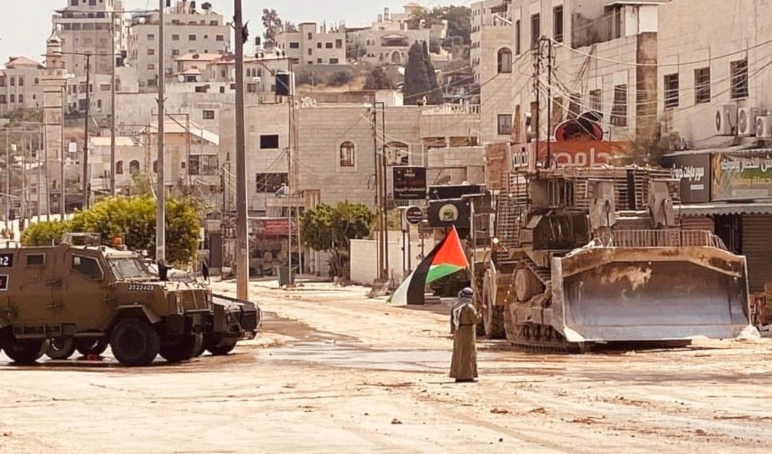 A Palestinian elder fearlessly holds up the Palestinian flag in front of invading Israeli occupation forces vehicles in Tulkarm, occupied West Bank, amid an ongoing 8 hours raid on August 22, 2024. (Social media)