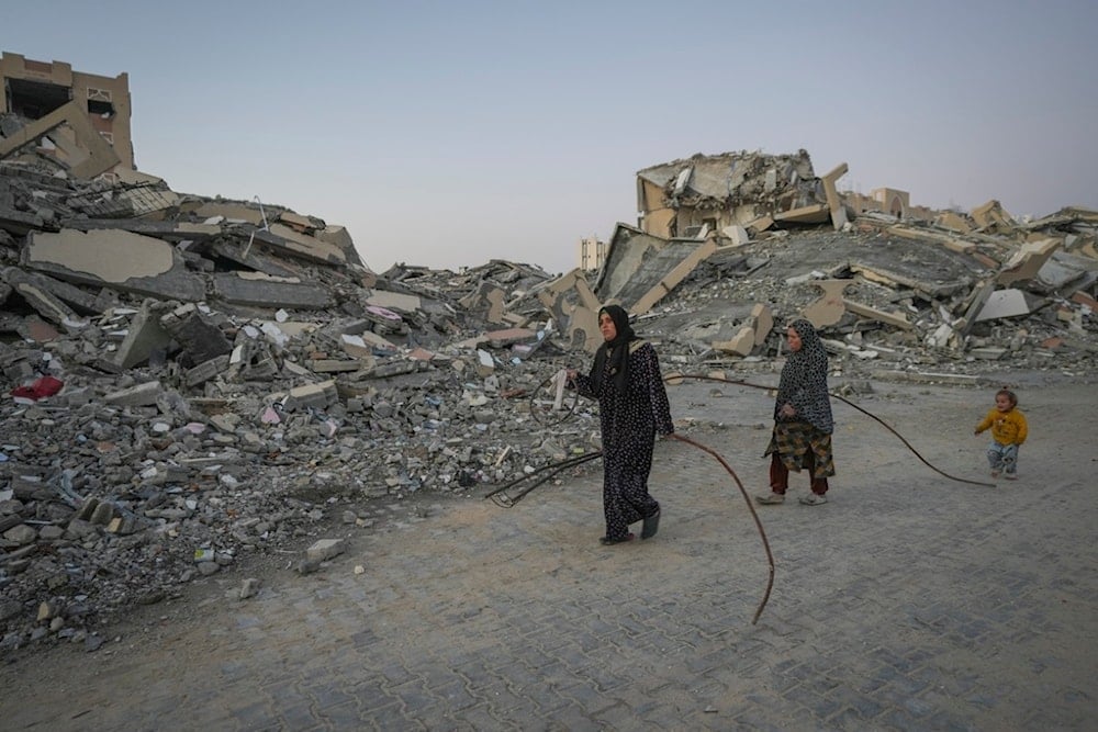 Followed by a young girl, two Palestinian women carry metal bars salvaged from the ruins to reinforce their tents at their camp in the central Gaza Strip town of Khan Younis, Wednesday, January 1, 2025 (AP)