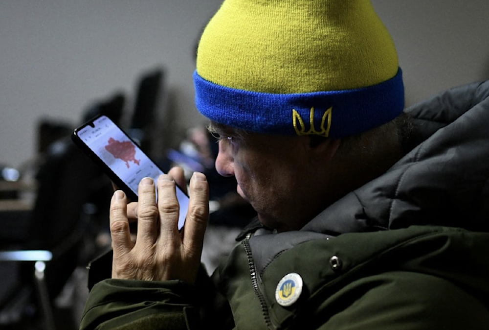 A Ukrainian man checks an air raid alert map on his phone while sitting in an underground hotel shelter during an air raid in Kyiv - November 25, 2025 (AFP)
