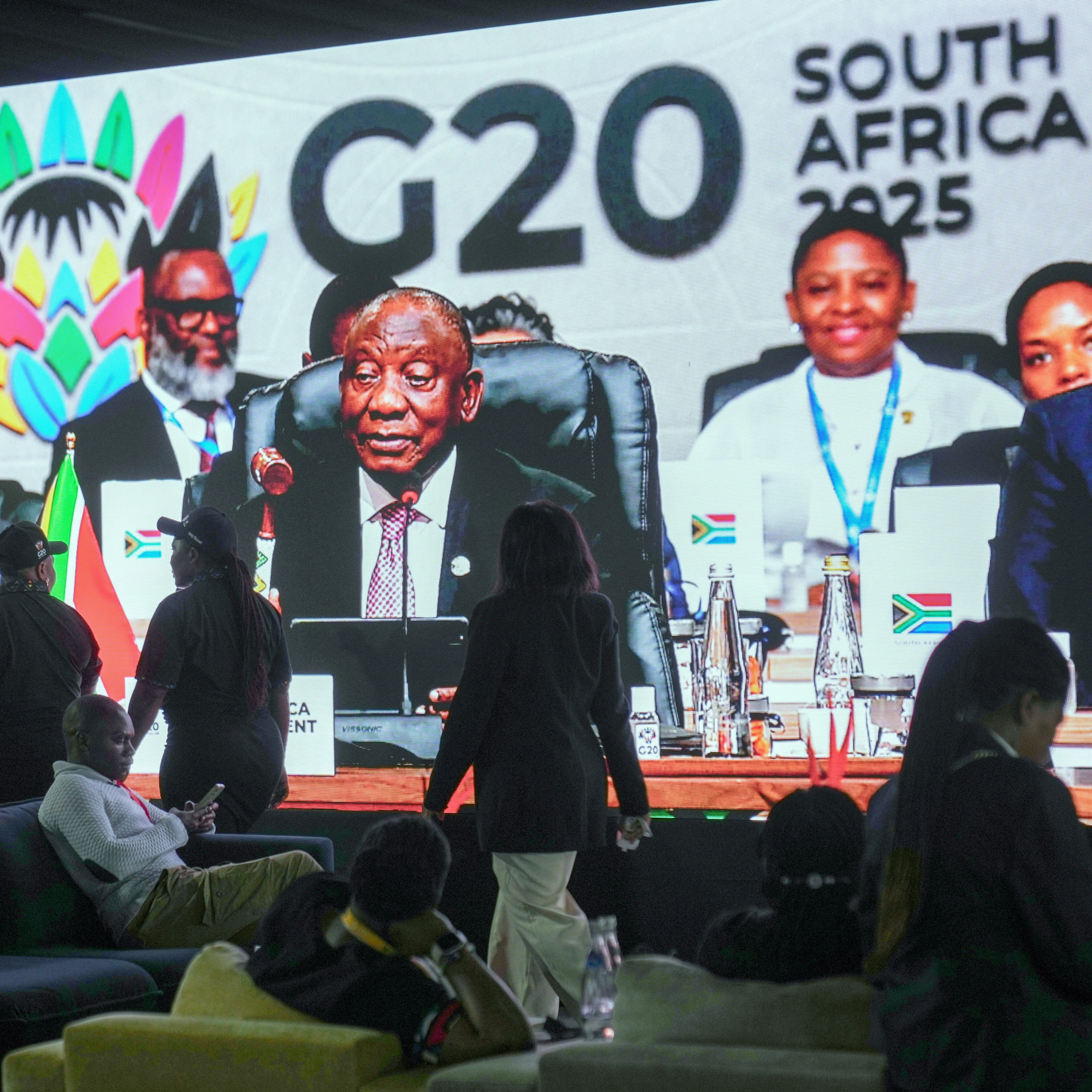 People walk by a large screen TV where South African President Cyril Ramaphosa holds a wooden gavel as he officially closes the G20 leaders' summit, in Johannesburg, South Africa, Sunday, Nov. 23, 2025 (AP)