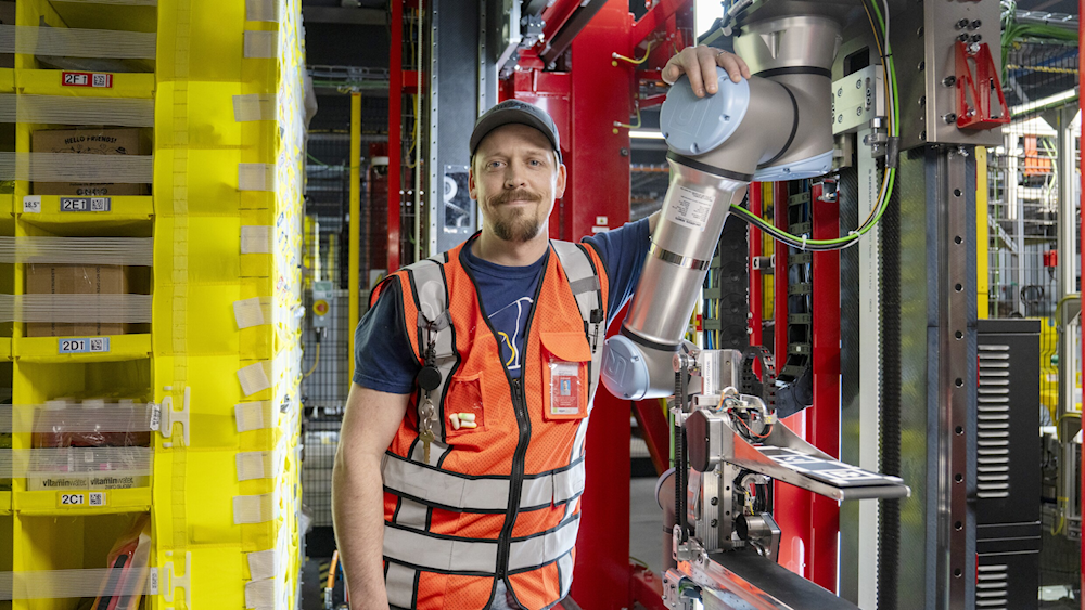 An Amazon manufacturing worker stands next to the Vulcan robot at an Amazon work site (Amazon website)