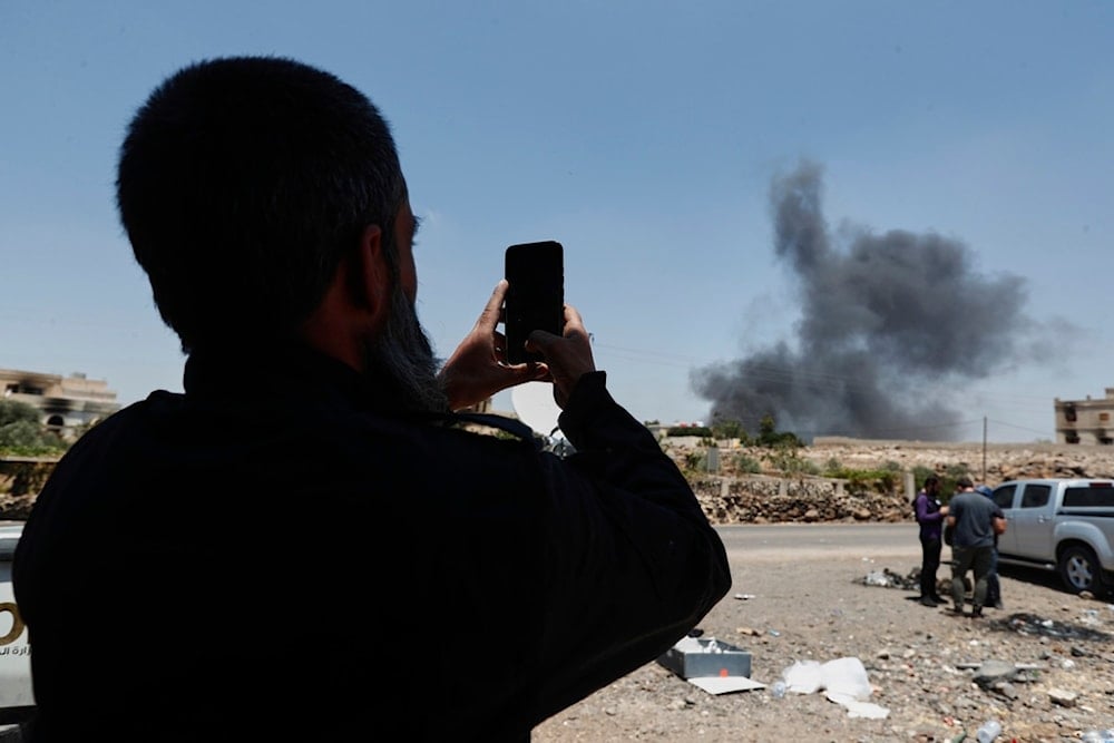 A Syrian security officer uses his phone to film smoke rising from burning houses in Mazraa village on the outskirts of Sweida city, in southern Syria, Sunday, July 20, 2025 (AP)