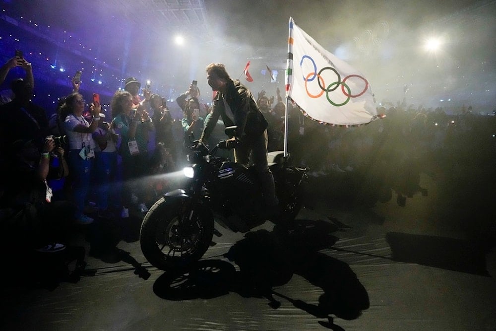 Tom Cruise rides a motorbike with the Olympic flag attached past athletes during the 2024 Summer Olympics closing ceremony at the Stade de France, Sunday, August 11, 2024, in Saint-Denis, France. (AP Photo/Ashley Landis)