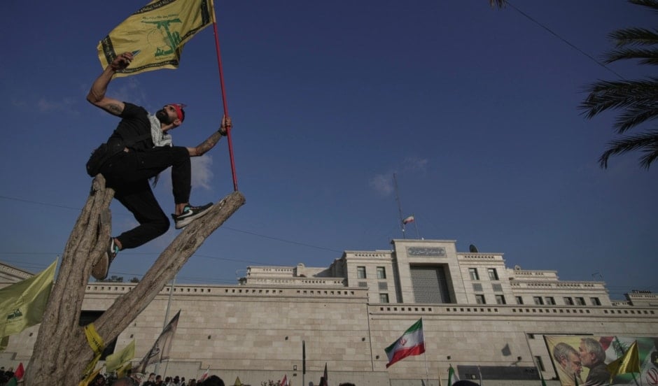 A Lebanese man takes a selfie while waving a Hezbollah flag as he stands on a tree outside the Iranian Embassy in Beirut, Lebanon, Wednesday, June 25, 2025 (AP)
